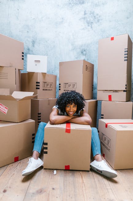 A young woman with curly dark hair is seated on a light wooden floor in a room with a light blue textured wall, surrounded by numerous cardboard boxes of various sizes, some sealed with red or black tape and others partially open. She is resting her arms on a large, closed cardboard box in front of her, smiling at the camera. The boxes are stacked and arranged around her, indicating a packing or home relocation process. The image captures the scene during a moving or packing stage, with the boxes prepared for transportation or storage as part of a household move supported by Croydon Removals. The setting suggests an indoor environment dedicated to packing and loading activities, with no visible furniture or appliances other than the boxes, emphasizing the focus on packing and careful handling during a furniture transport or moving service.