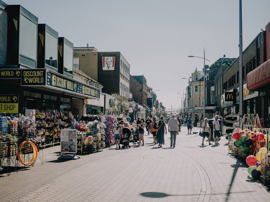 The image depicts a busy outdoor shopping street with various retail stores, including Discount World, a McDonald's, and other small shops along a paved pedestrian area. On the left side, shopfronts display merchandise such as flowers, balloons, and outdoor display racks, with some items on carts and tables. The right side features additional small shops and storefronts, with street lamps and awnings providing shade. Several pedestrians, some pushing strollers or carrying shopping bags, walk along the street, while others browse the displayed goods outside the stores. The weather appears sunny with a clear sky, and the scene captures a lively commercial environment typical of a town centre. This setting could relate to house removals or packing activities, reflecting the importance of logistics and transportation in local business relocations, as handled by Croydon Removals.
