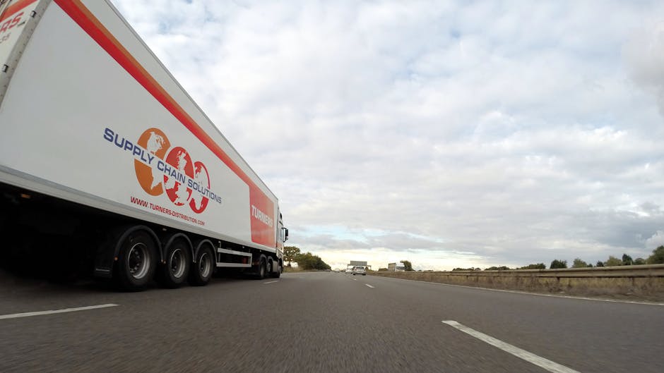 A large white moving truck with the logo and branding of Turners Distribution on the side, featuring the phrase 'Supply Chain Solutions' and the website URL, is situated on a multi-lane road, part of a home relocation or furniture transport process. The truck is positioned on the left side of the image, with its rear section visible and the cab extending towards the right. The scene appears to be outdoors under a cloudy sky, with other vehicles visible in the distance. The truck is parked or moving along a road with a guardrail on the right, typical of a highway or major road suitable for commercial deliveries or relocations. The setting suggests an active loading or transit phase, relevant to professional removals services provided by Croydon Removals, as part of their packing and moving logistics for local businesses or household moves in Croydon, depicted in the page titled 'Purley Way Commercial Removals for local businesses, Croydon'.