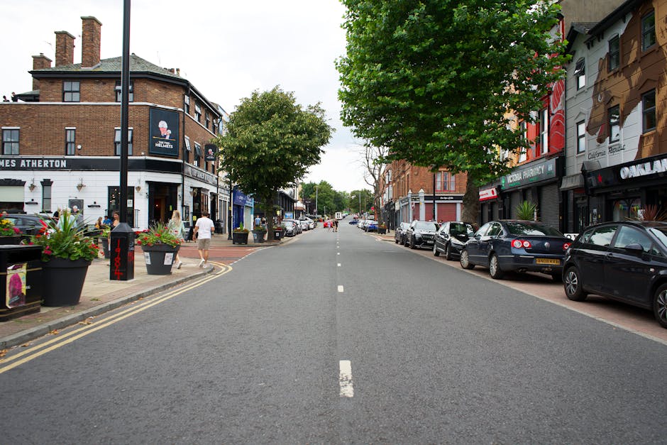 The image depicts a busy outdoor shopping street with various retail stores, including Discount World, a McDonald's, and other small shops along a paved pedestrian area. On the left side, shopfronts display merchandise such as flowers, balloons, and outdoor display racks, with some items on carts and tables. The right side features additional small shops and storefronts, with street lamps and awnings providing shade. Several pedestrians, some pushing strollers or carrying shopping bags, walk along the street, while others browse the displayed goods outside the stores. The weather appears sunny with a clear sky, and the scene captures a lively commercial environment typical of a town centre. This setting could relate to house removals or packing activities, reflecting the importance of logistics and transportation in local business relocations, as handled by Croydon Removals.