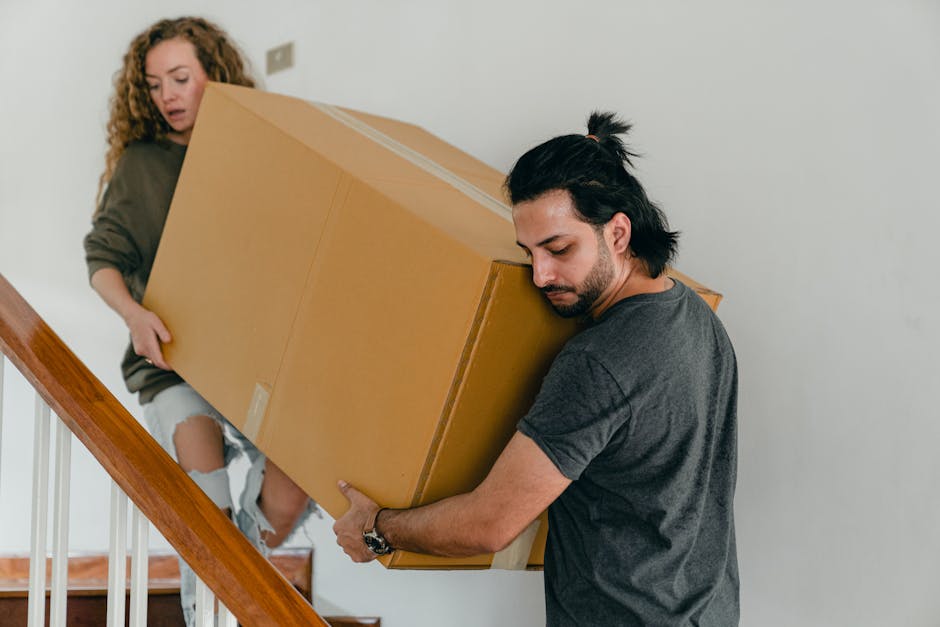 A man with dark hair tied back and wearing a dark grey t-shirt is carefully carrying a large cardboard box up a staircase, supporting it with both hands. Behind him, a woman with curly hair, dressed in a brown sweater and ripped jeans, is also holding the box from the other side, assisting with the move. The staircase has wooden steps and white railings, and the interior wall is plain white. The scene depicts a home removal or furniture transport process as part of a packing and moving operation, possibly organized by Croydon Removals, in a residential property. The lighting appears natural, highlighting the details of the packing materials and the coordinated effort involved in the moving process. The image captures a moment of physical loading during a house relocation, emphasizing the importance of careful handling of boxes and furniture during a professional removals service.