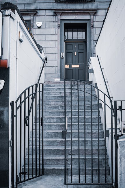A set of concrete stairs leading up to a dark grey door with a window grid, situated on the front of a grey stone building. The stairs are flanked by white walls on either side, with a black wrought iron gate at the base, partially open. To the left of the stairs, there is a small outdoor light fixture mounted on the wall. A dispatch card or notice is visible on the door. The entrance area appears to be part of a home or apartment in a city environment, with an urban facade. The overall scene captures a typical stairway leading to a residential door, relevant to home relocation and furniture transport services offered by Croydon Removals, demonstrating typical access features used during house removals or move-in processes.