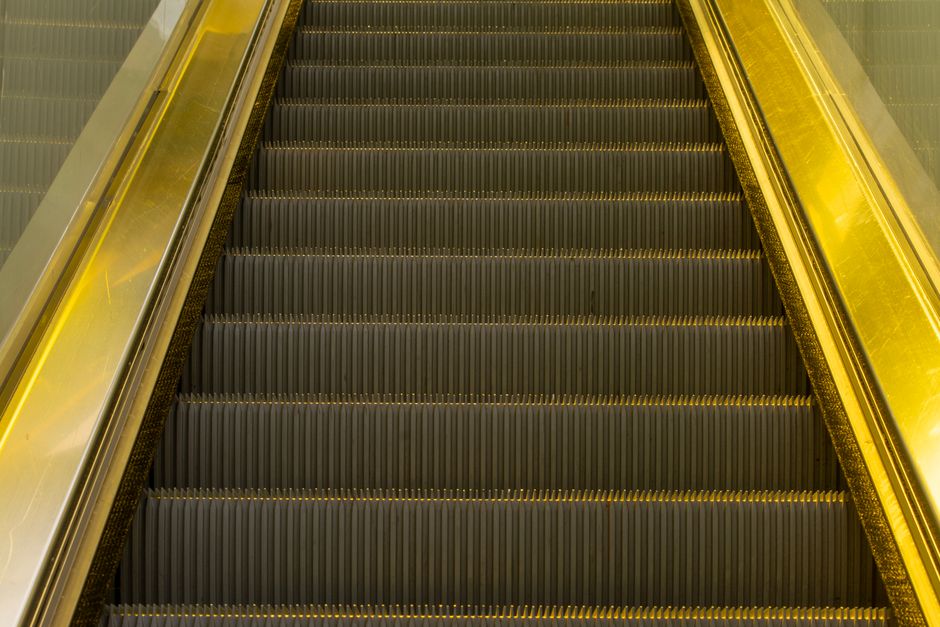 A man with dark hair tied back and wearing a dark grey t-shirt is carefully carrying a large cardboard box up a staircase, supporting it with both hands. Behind him, a woman with curly hair, dressed in a brown sweater and ripped jeans, is also holding the box from the other side, assisting with the move. The staircase has wooden steps and white railings, and the interior wall is plain white. The scene depicts a home removal or furniture transport process as part of a packing and moving operation, possibly organized by Croydon Removals, in a residential property. The lighting appears natural, highlighting the details of the packing materials and the coordinated effort involved in the moving process. The image captures a moment of physical loading during a house relocation, emphasizing the importance of careful handling of boxes and furniture during a professional removals service.